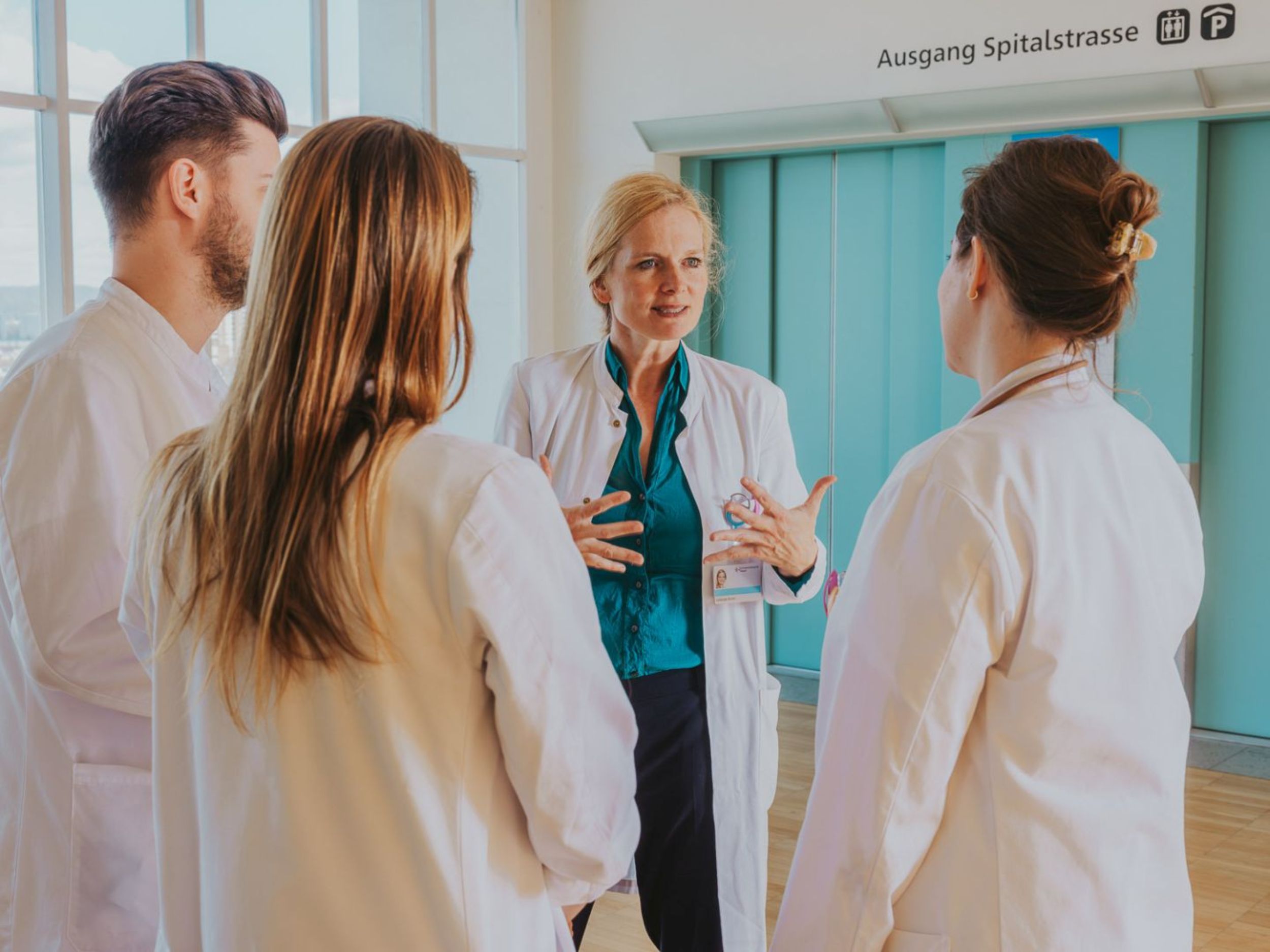Eine Ärztin im weissen Kittel spricht gestikulierend mit drei Mitarbeitenden und führt ein Gespräch im Team in einem hellen Krankenhausbereich.
