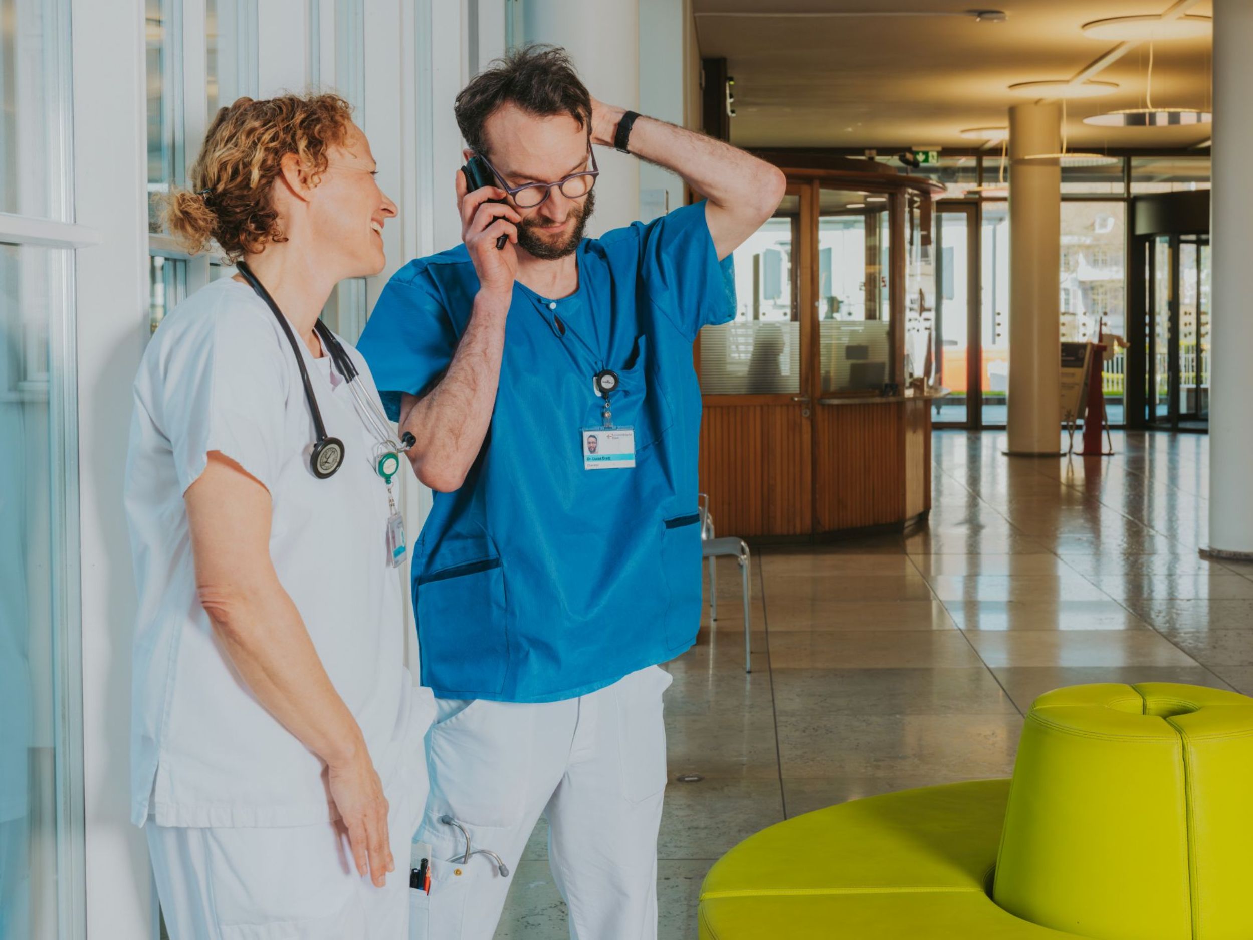 Ärztin und Arzt stehen im Krankenhausflur, unterhalten sich und telefonieren gleichzeitig; beide wirken entspannt in einer kurzen Arbeitspause.