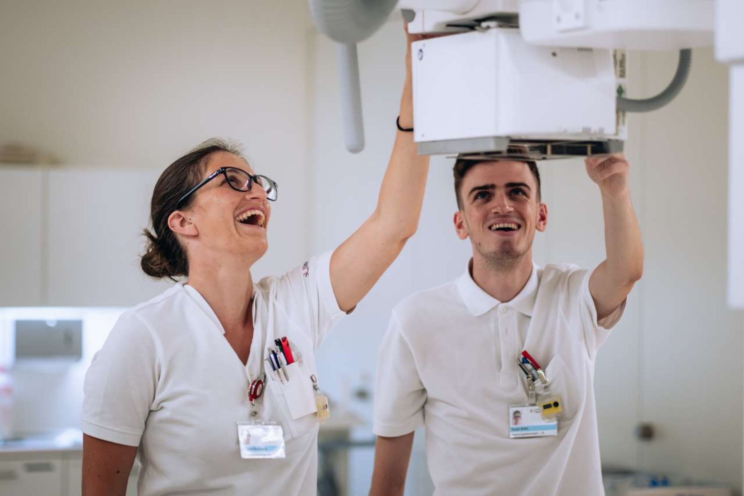Zwei Radiologiefachpersonen in weisser Berufskleidung bedienen gemeinsam ein Röntgengerät und lachen.