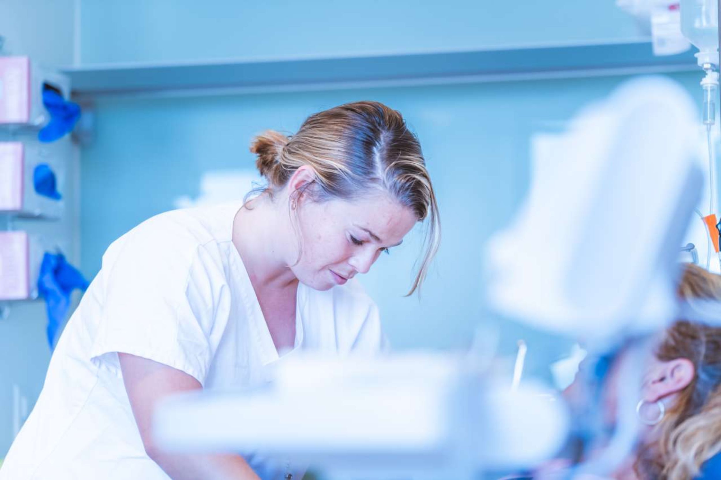 Eine Frau in weisser Berufskleidung beugt sich konzentriert über eine Patientin in einer medizinischen Umgebung. Der Hintergrund zeigt Wandregale mit blauen Handschuhen und anderen medizinischen Utensilien. Die Szene zeigt eine Pflege- oder Behandlungssituation in einer professionellen medizinischen Einrichtung.