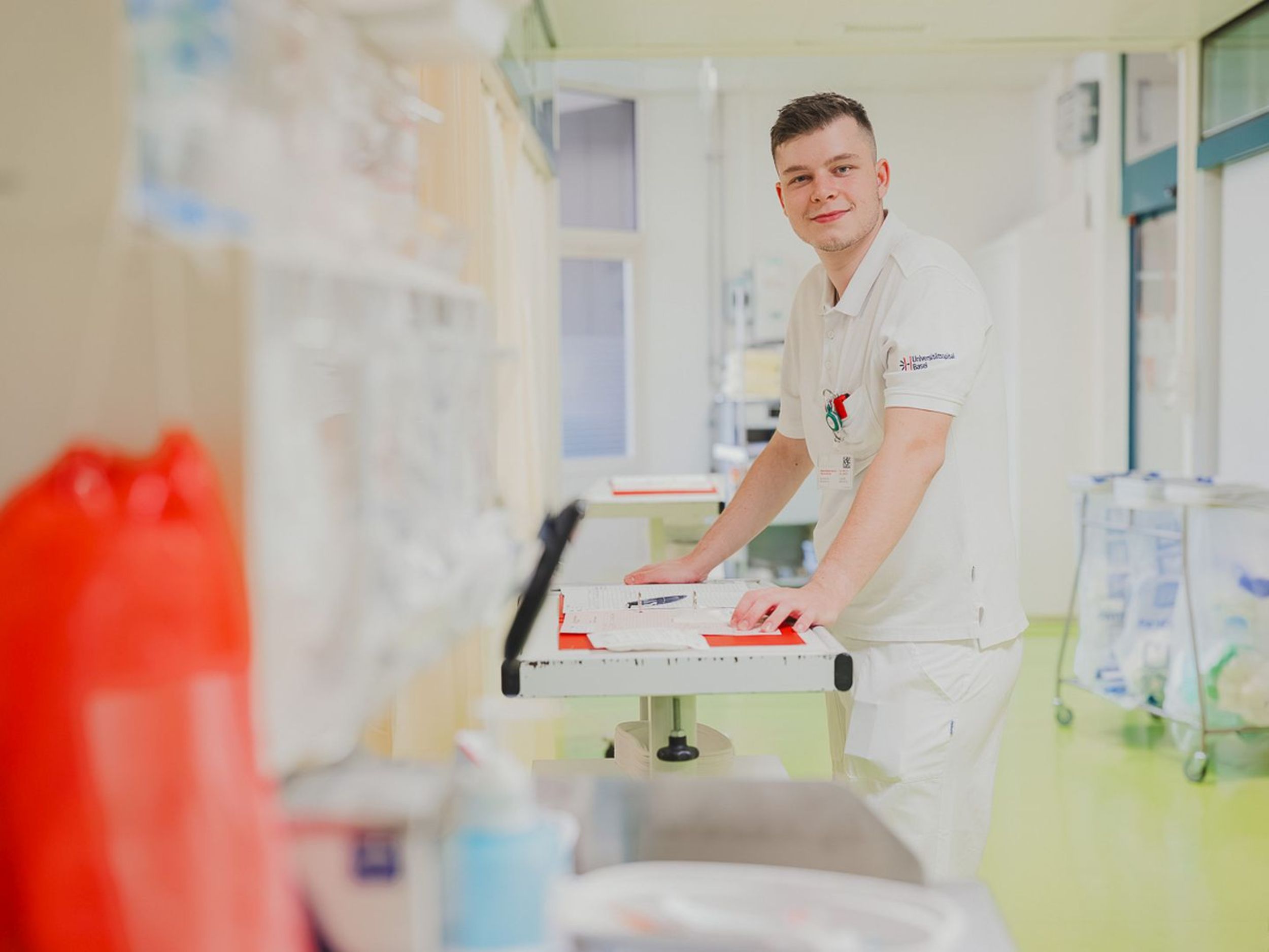 Eine Szene in einer Spitalumgebung zeigt medizinisches Personal in weisser Berufskleidung, das sich um einen Patienten auf einem Bett kümmert. Ein Mann schiebt das Bett durch einen Korridor, während eine weitere Person mit einer Maske das Bett begleitet. Im Vordergrund steht ein weiterer Mitarbeiter an einem Tisch. Die Umgebung ist klar strukturiert, mit medizinischen Geräten, Monitoren und Aufbewahrungssystemen. Die Szene zeigt eine Intensiv- bzw. Notfallstation.