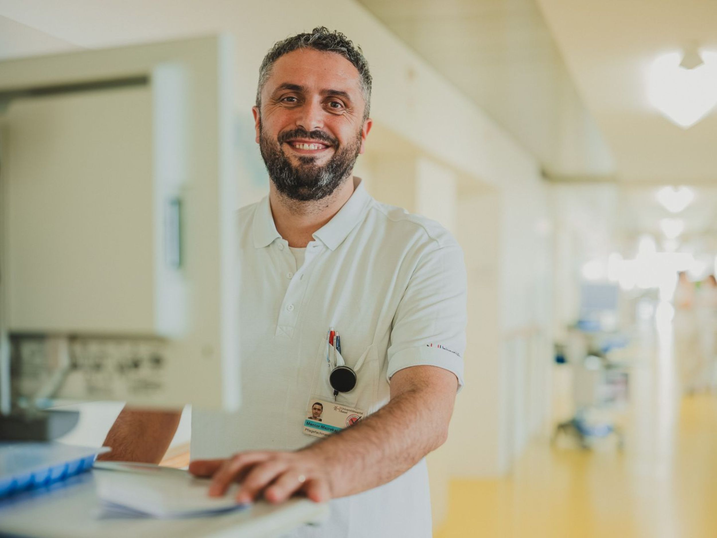Ein lächelnder Pflegefachmann in weisser Arbeitskleidung steht an einem Tresen im Krankenhausflur und bedient einen Computer.