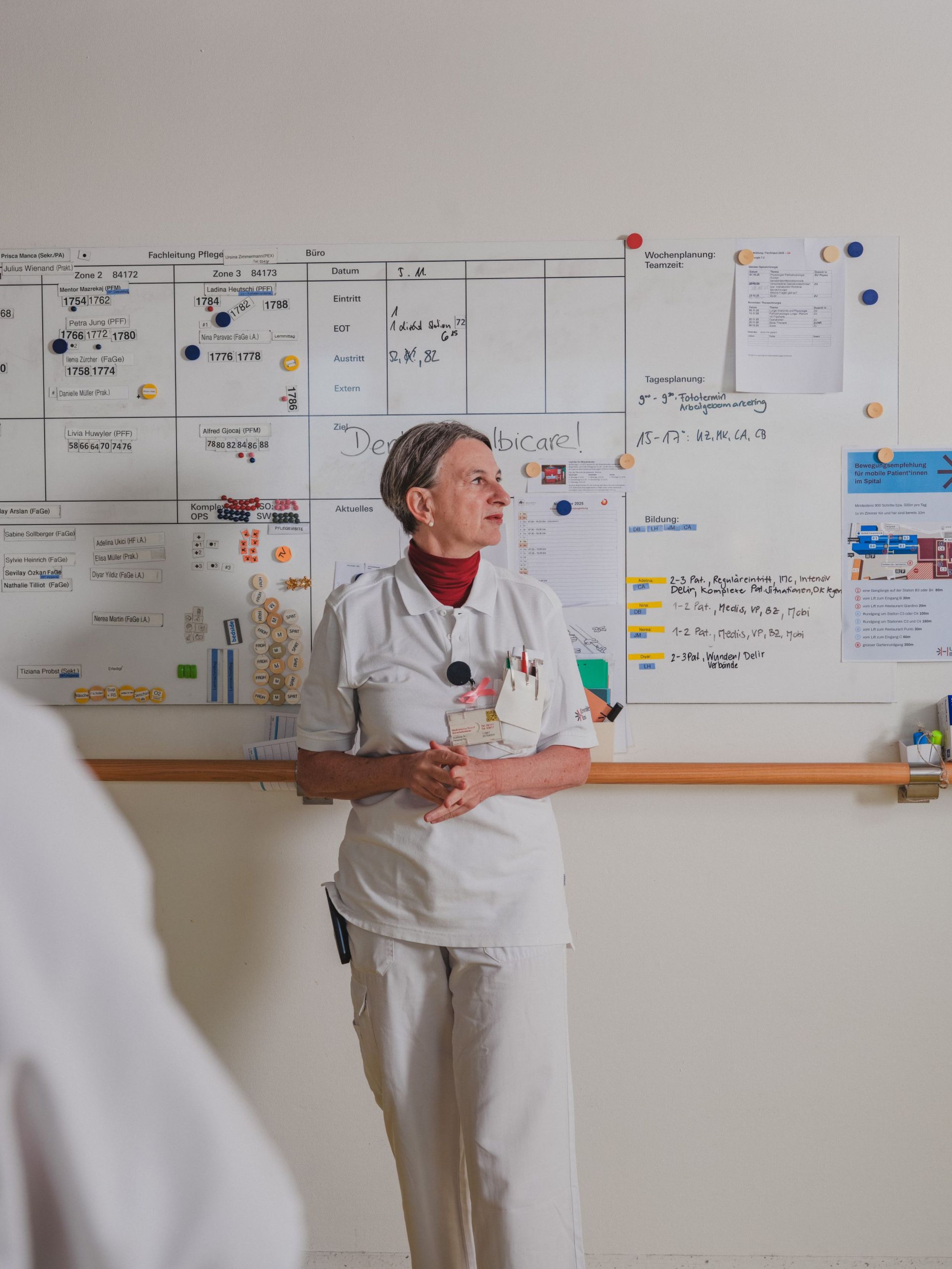 Eine Pflegefachfrau in weisser Arbeitskleidung steht vor einem Whiteboard mit Dienstplanung und Informationen und spricht im Rahmen einer Teambesprechung.