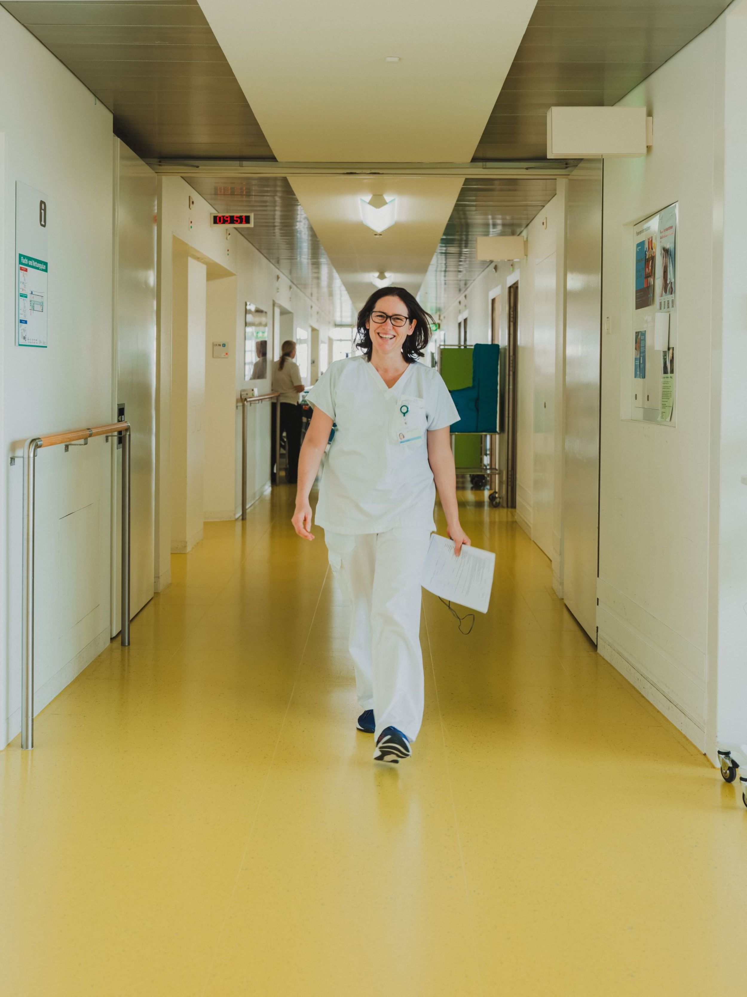 Eine Pflegekraft in weisser Arbeitskleidung läuft lächelnd durch einen Krankenhausflur und hält ein Blatt Papier in der Hand.