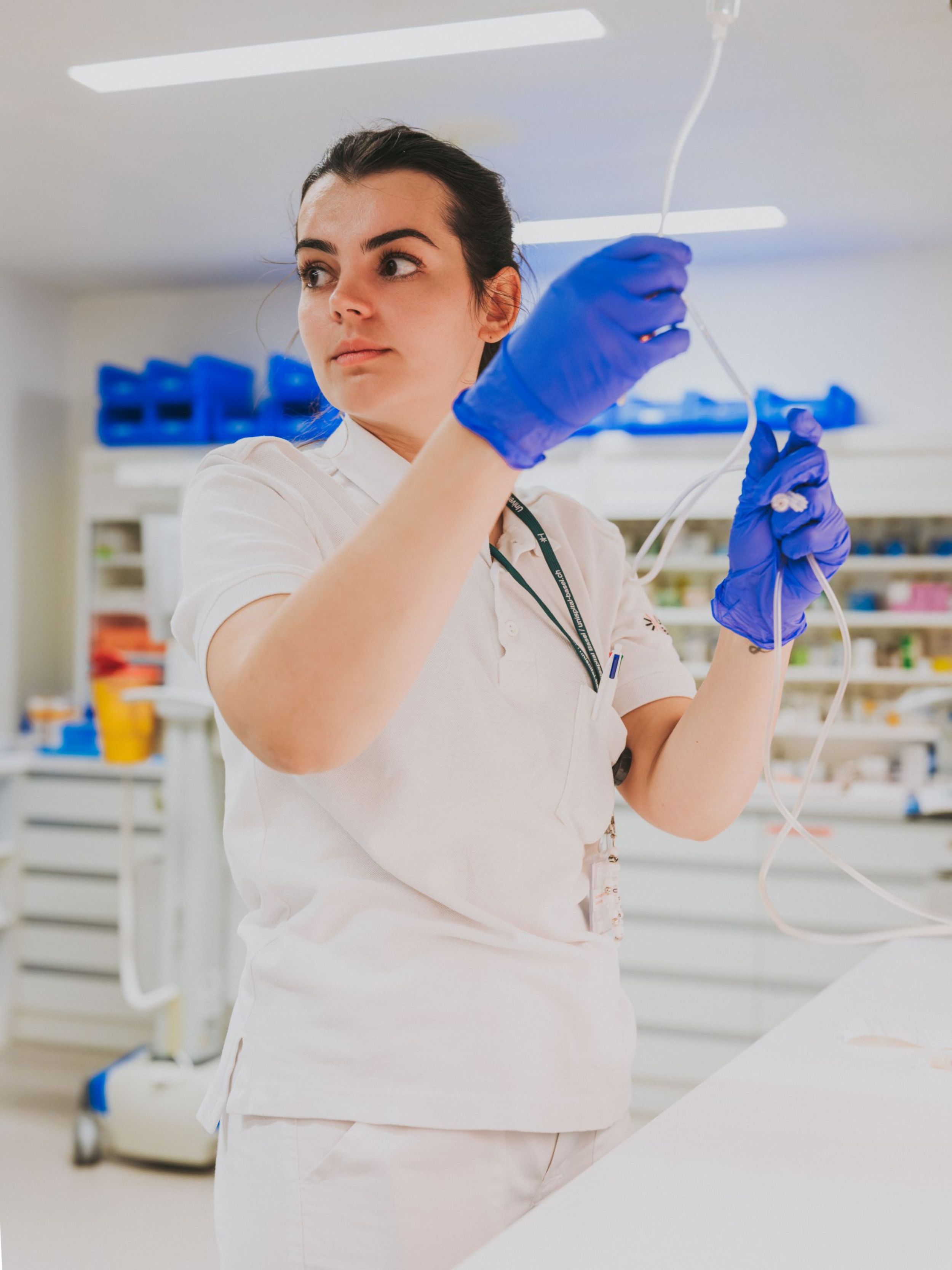 Pflegefachperson mit Handschuhen bereitet eine Infusion vor und hält den Schlauch prüfend hoch; im Hintergrund sind medizinische Materialien und ein Behandlungsraum zu sehen.