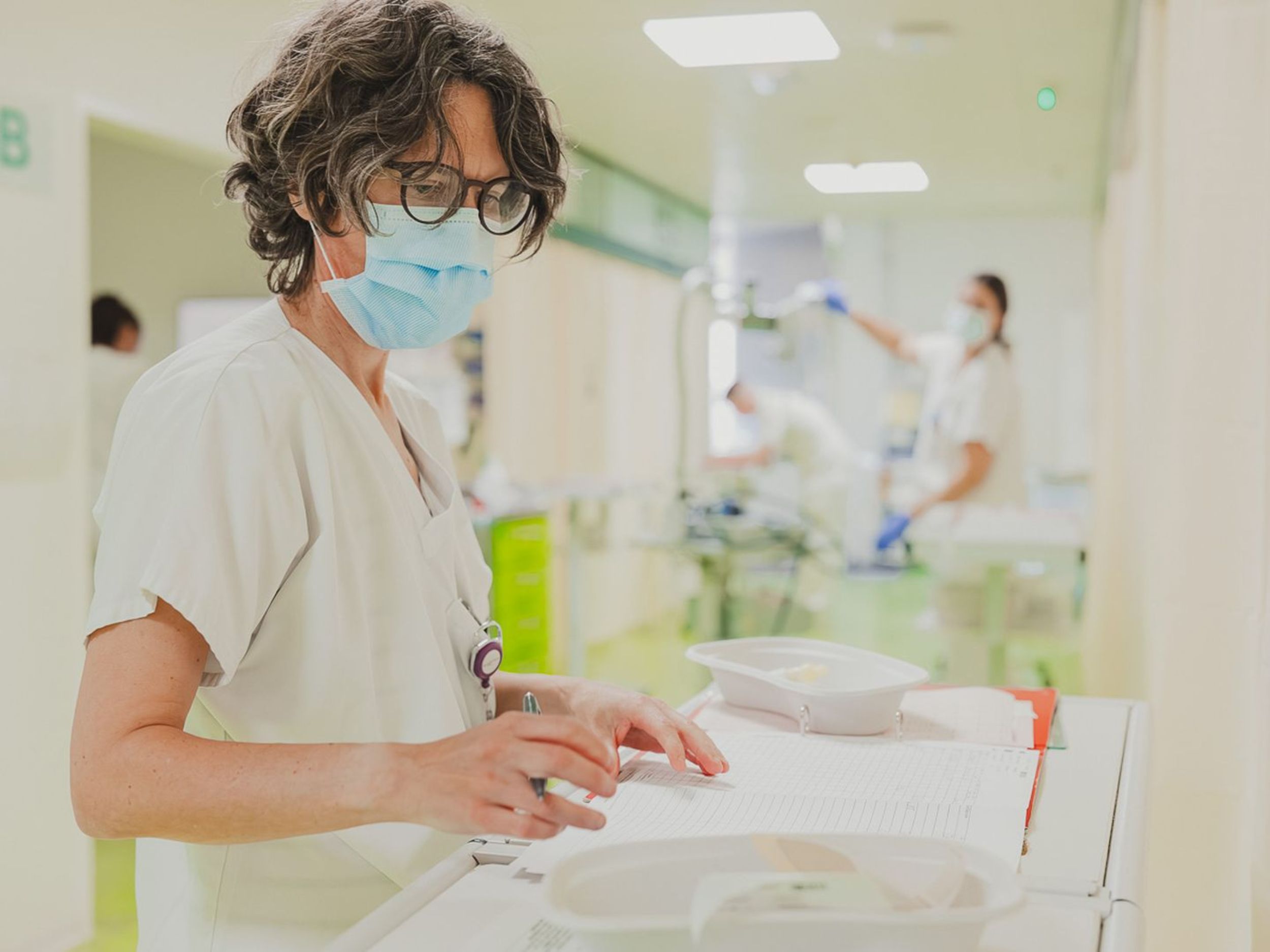 Eine Pflegefachfrau mit Brille und Mundschutz dokumentiert auf einem Klemmbrett, während sie an einem Pflegewagen im Krankenhausflur steht; im Hintergrund arbeitet eine weitere Pflegekraft.