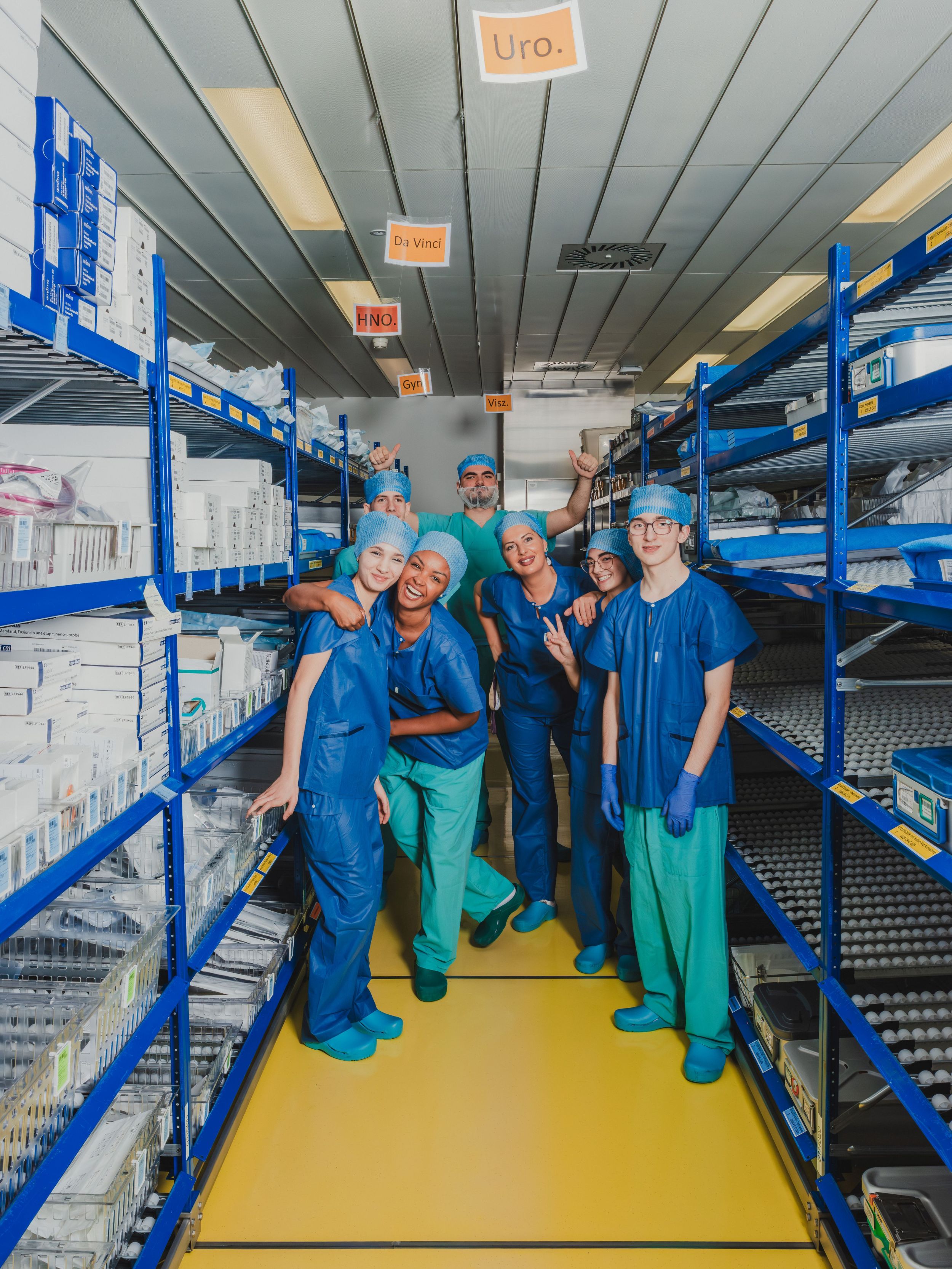 Gruppe von medizinischen Mitarbeitenden in Arbeitskleidung steht lächelnd zwischen Regalen mit steril verpackten Materialien in einem Lagerraum; über ihnen hängen Beschriftungen für verschiedene Fachbereiche.