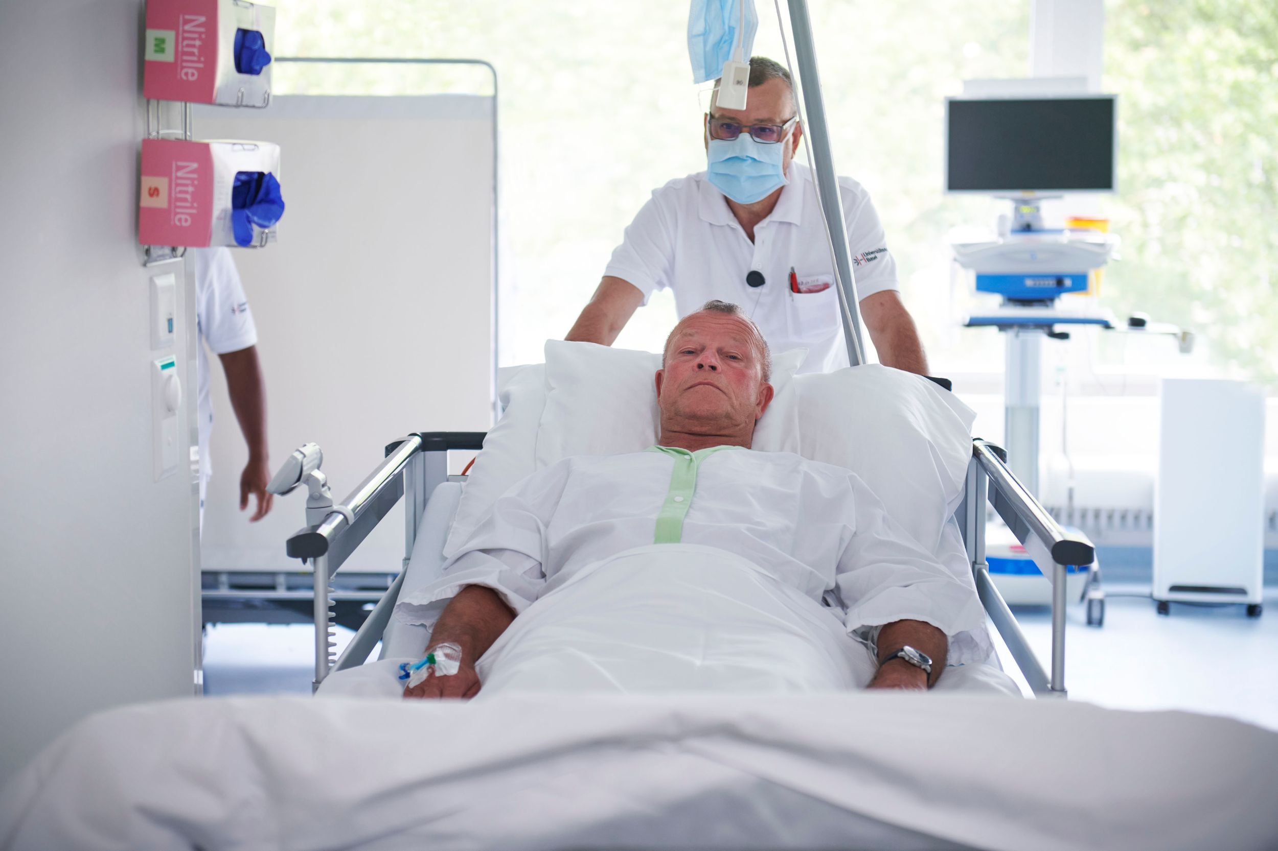 Patient wird im Bett von einem Pflegenden in den Gang geschoben