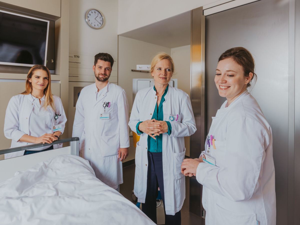 Vier medizinische Fachpersonen in weissen Kitteln stehen gemeinsam in einem Patientenzimmer neben einem Spitalbett und lächeln; die Situation wirkt wie ein Teamgespräch oder eine Visite.