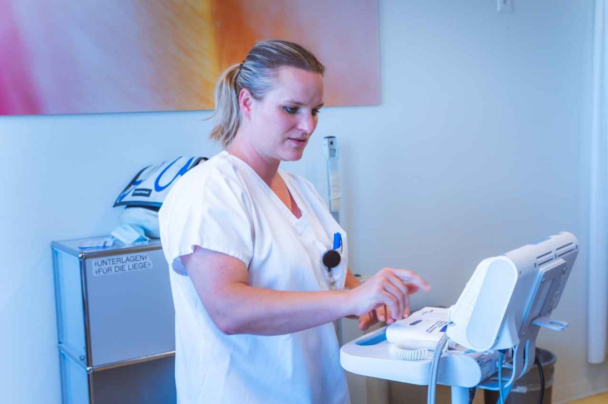 Eine Frau in weisser Berufskleidung steht in einem medizinischen Raum und bedient ein elektronisches Gerät mit mehreren Kabeln und Bildschirmen. Sie wirkt konzentriert, während sie eine Taste drückt. Im Hintergrund sind ein Schrank sowie ein buntes abstraktes Bild an der Wand zu sehen. Die Umgebung zeigt einen professionellen medizinischen Arbeitsplatz.