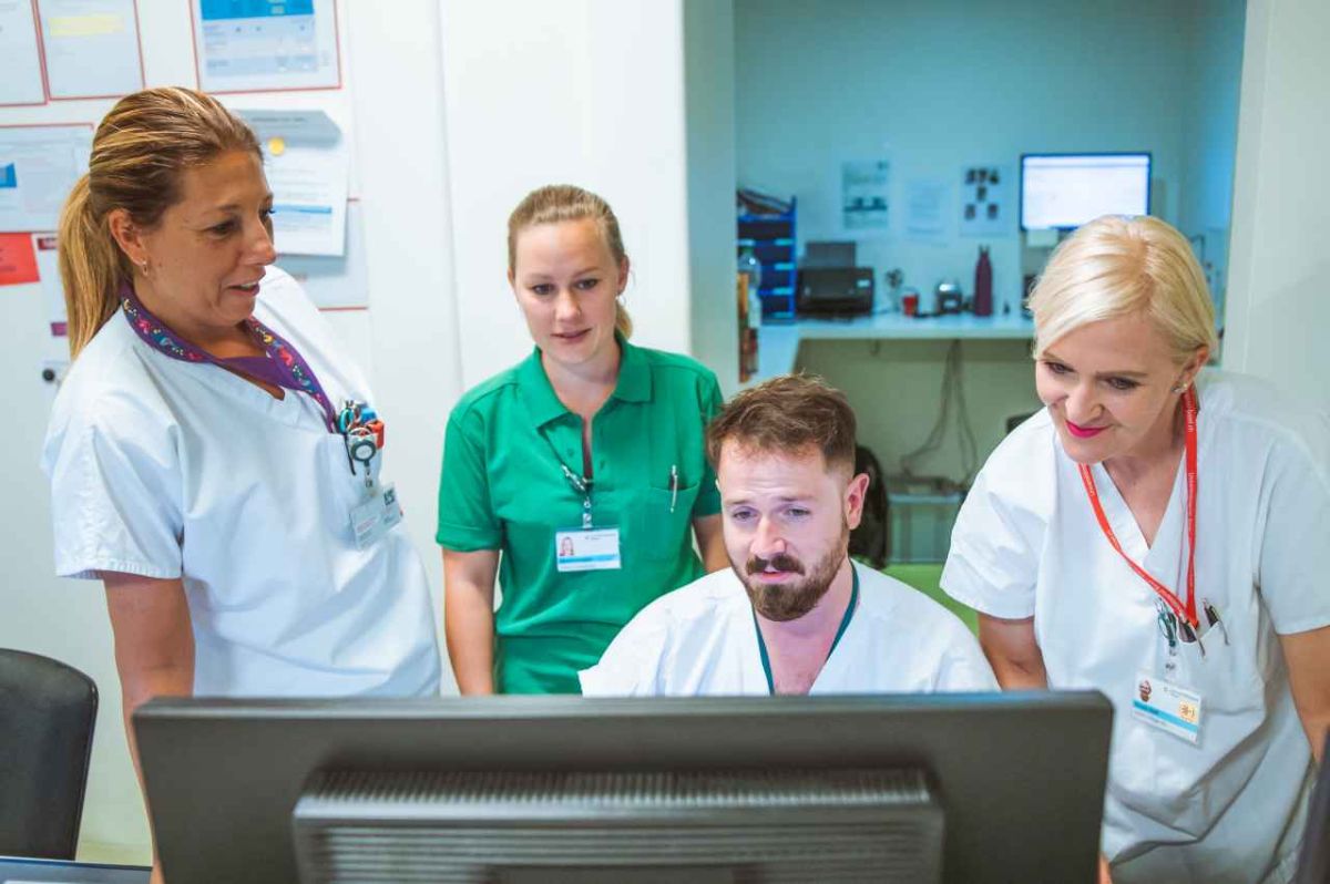 Vier Personen, drei Frauen und ein Mann, stehen oder sitzen um einen Computerbildschirm in einer Arbeitsumgebung. Die Frauen tragen weisse und grüne Berufskleidung, der Mann ebenfalls Weiss. Sie blicken konzentriert auf den Bildschirm, während sie möglicherweise etwas besprechen oder überprüfen. Im Hintergrund sind Wände mit Dokumenten und ein weiterer Computerarbeitsplatz sichtbar. Die Szene zeigt eine gemeinsame, berufsbezogene Tätigkeit im medizinischen Bereich.