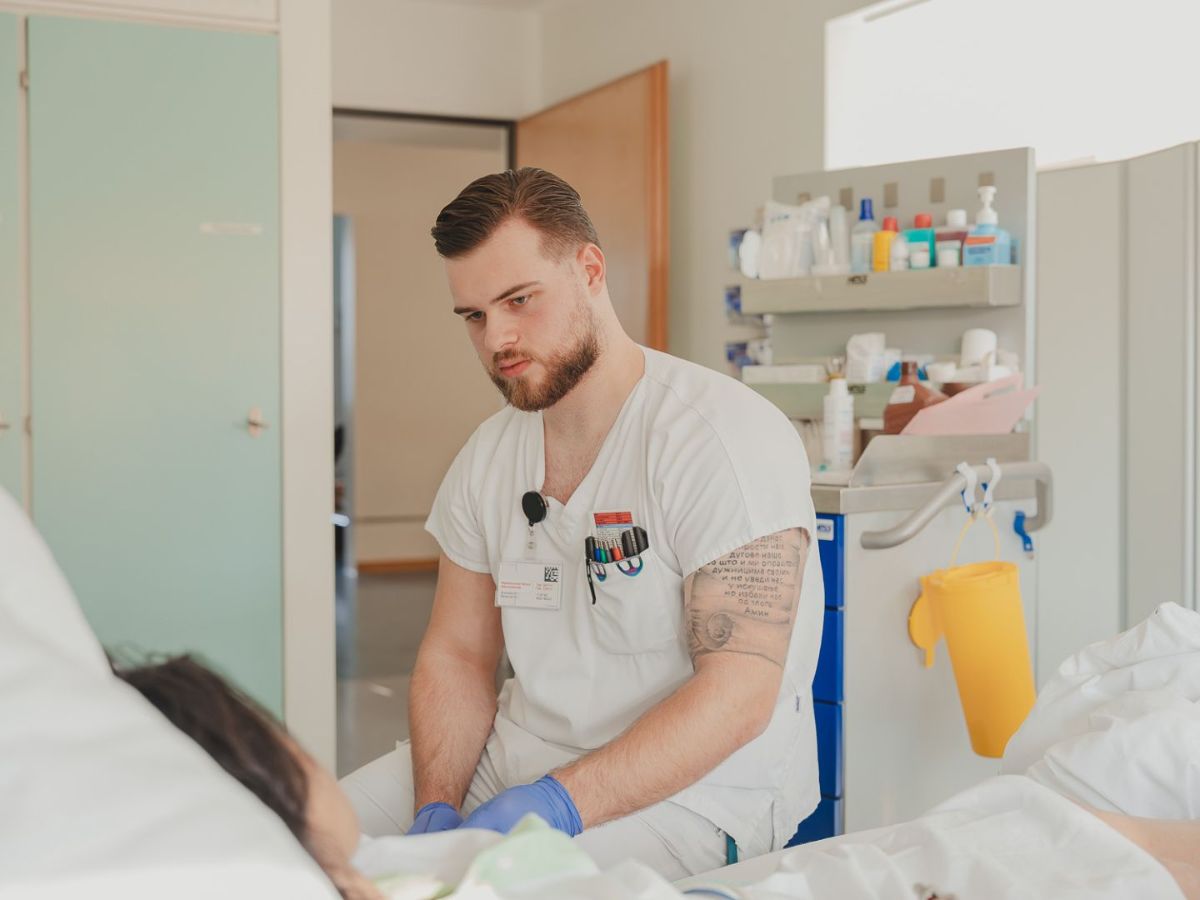 Ein Pflegefachmann in weisser Arbeitskleidung versorgt eine Patientin im Krankenhausbett und führt dabei eine Behandlung mit Handschuhen durch.