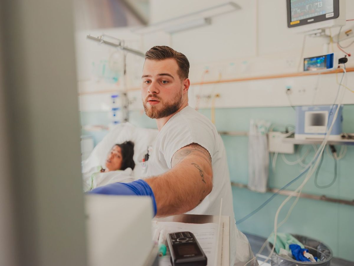 Ein Pflegefachmann in weisser Kleidung bedient ein medizinisches Gerät am Bett, während im Hintergrund eine Patientin im Krankenhausbett liegt.
