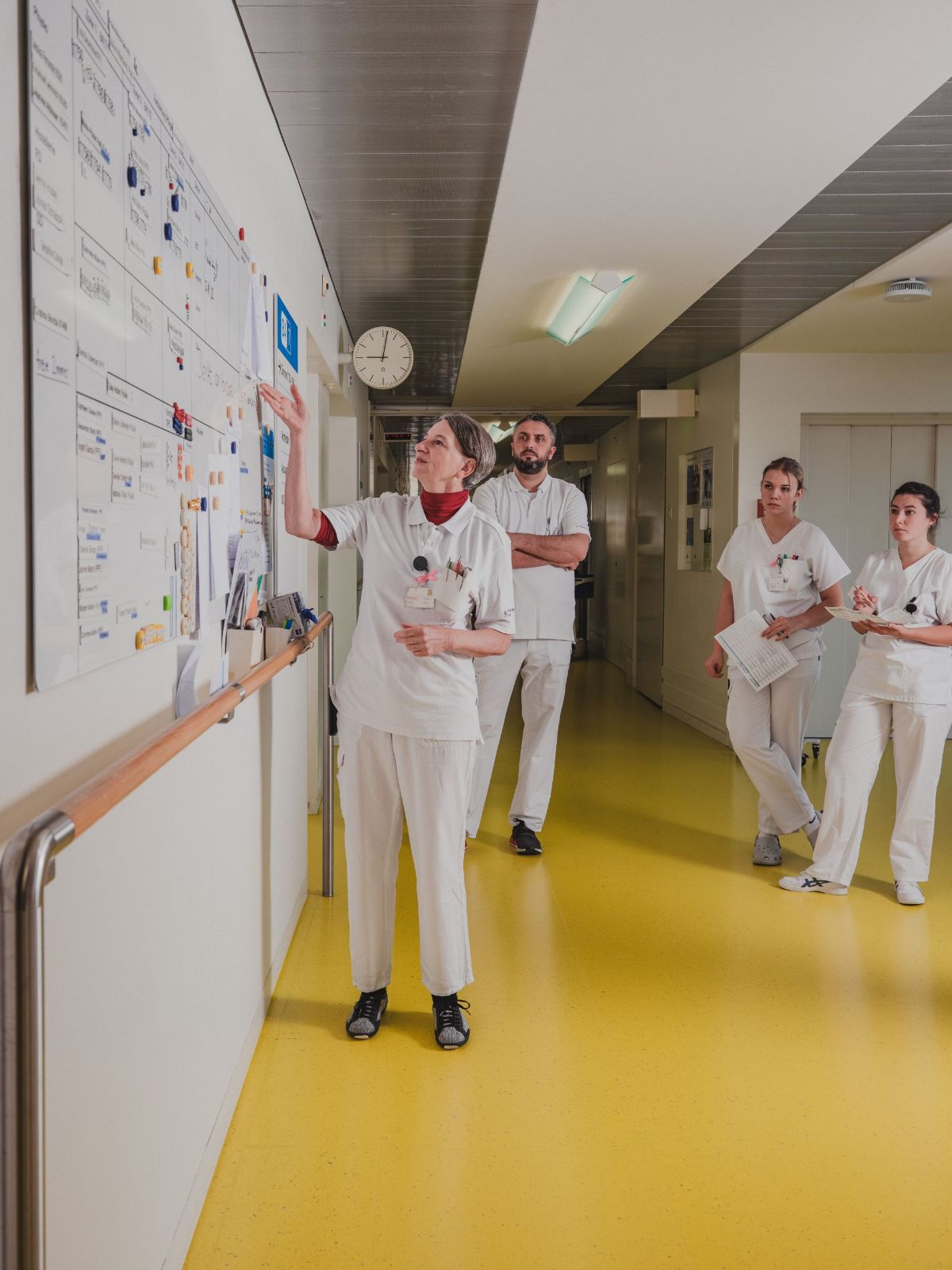 Eine Pflegekraft zeigt auf einen Dienstplan an der Wand eines Krankenhausflurs, während drei weitere Mitarbeitende in weisser Kleidung aufmerksam zusehen und Notizen halten.