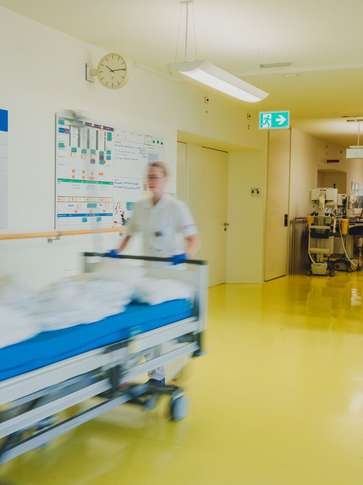 Pflegefachperson schiebt ein Spitalbett zügig durch einen hellen Krankenhausflur; an der Wand sind ein Einsatzplan, eine Uhr und ein Notausgangsschild zu sehen.
