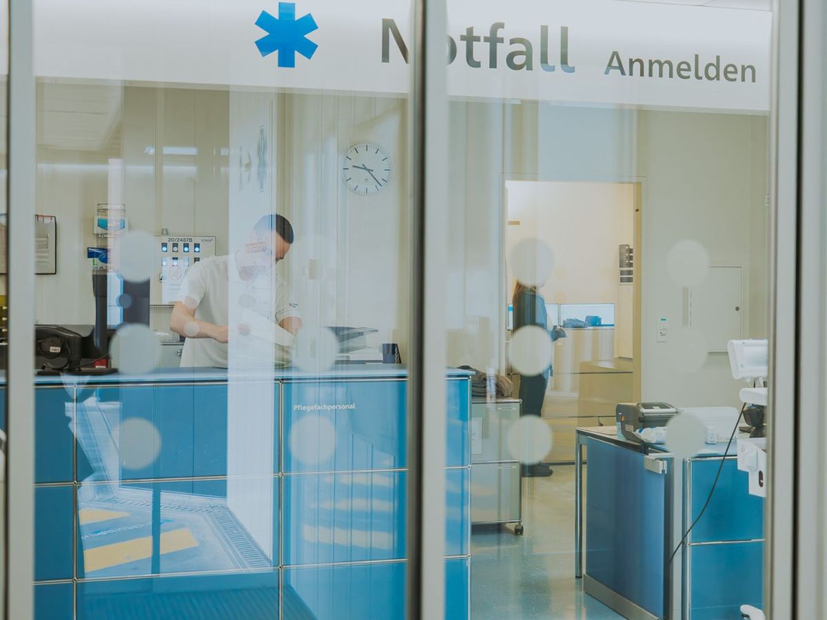 Pflegefachpersonen in weissen Uniformen und mit Gesichtsmasken stehen in einer Gruppe vor einem Whiteboard in einem Spital. Eine Frau zeigt mit einem Stift auf das Board und erklärt etwas, während die anderen aufmerksam zuhören. Die Szene wirkt professionell und konzentriert, typischerweise im Rahmen einer Team- oder Übergabebesprechung.