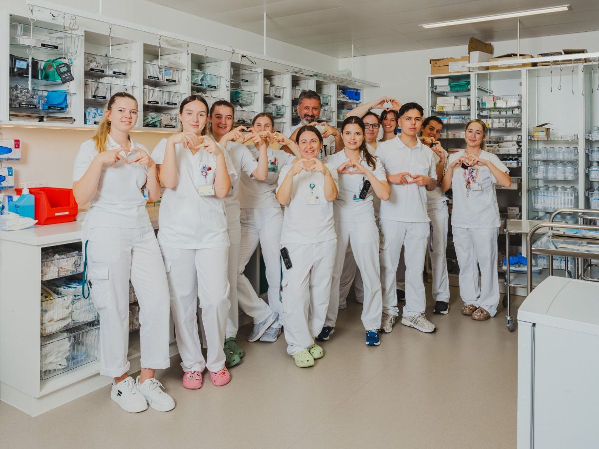 Pflegefachpersonen in weisser Arbeitskleidung stehen gemeinsam in einem Klinikraum, formen mit den Händen Herzsymbole und lächeln in die Kamera.