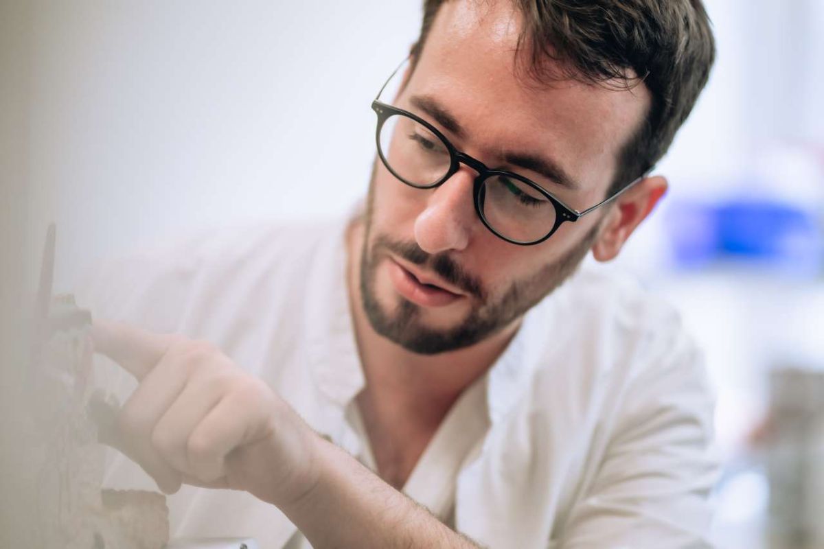 Ein Arzt mit Brille und weissem Kittel betrachtet konzentriert ein medizinisches Modell und zeigt mit dem Finger auf ein Detail. Die Szene vermittelt Fachwissen, Präzision und Engagement im klinischen oder forschungsnahen Arbeitsumfeld.