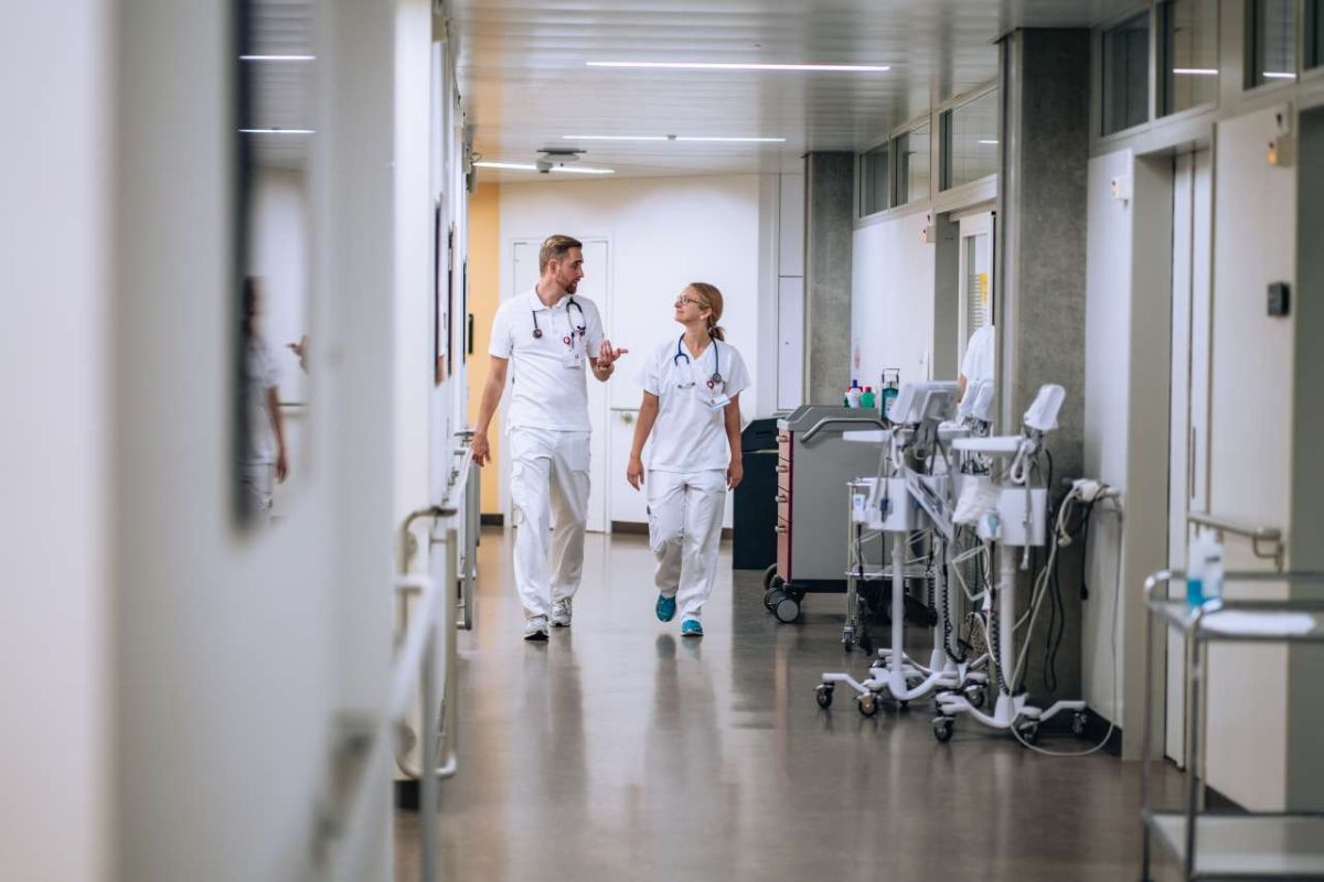 Ein Assistenzarzt und eine Assistenzärztin in weisser Berufskleidung gehen gemeinsam durch einen Spitalgang und unterhalten sich. Beide tragen ein Stethoskop – die Szene vermittelt Austausch, Teamarbeit und den ärztlichen Alltag im Spital.