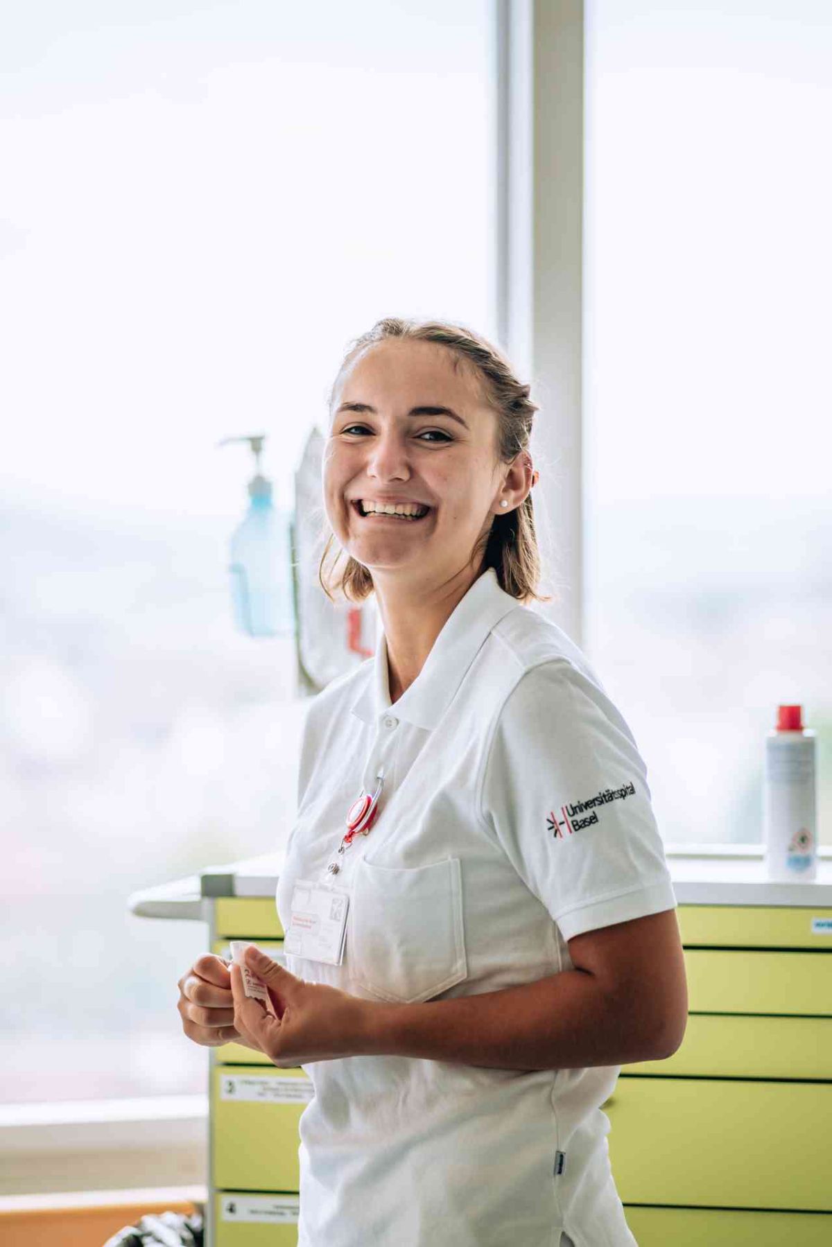 Eine junge Frau in weisser Berufskleidung steht lächelnd in einem hellen Raum. Sie trägt ein Namensschild mit der Aufschrift 'Universitätsspital Basel' auf ihrem Hemdärmel. Im Hintergrund sind ein Fenster, eine hellgrüne Schubladenkommode und medizinische Utensilien wie Desinfektionsmittel sichtbar. Die freundliche Ausstrahlung und das professionelle Umfeld vermitteln eine positive Arbeitsatmosphäre.