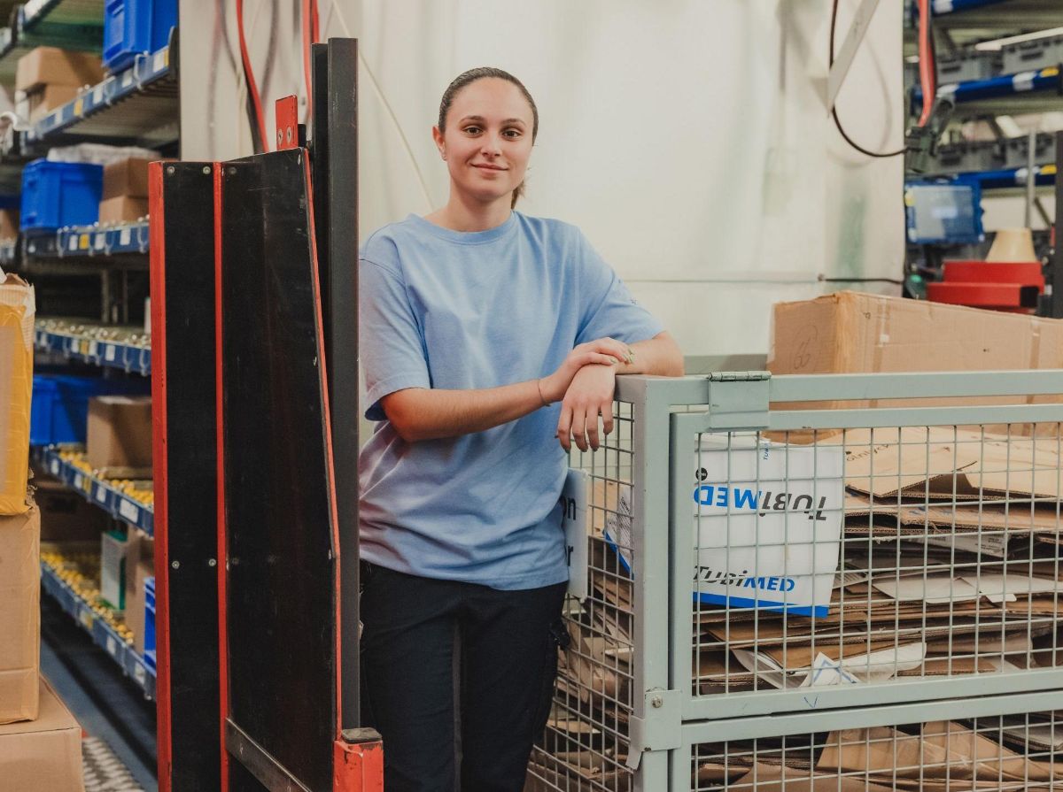 Zwei Lernende in hellblauen Arbeitsshirts stehen lächelnd in einem Lagerbereich mit Regalen und blauen Transportboxen.