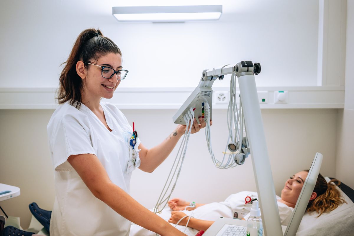 Medizinische Praxisassistentin in Ausbildung steht bei einer Patientin am Bett und bedient ein medizinisches Gerät