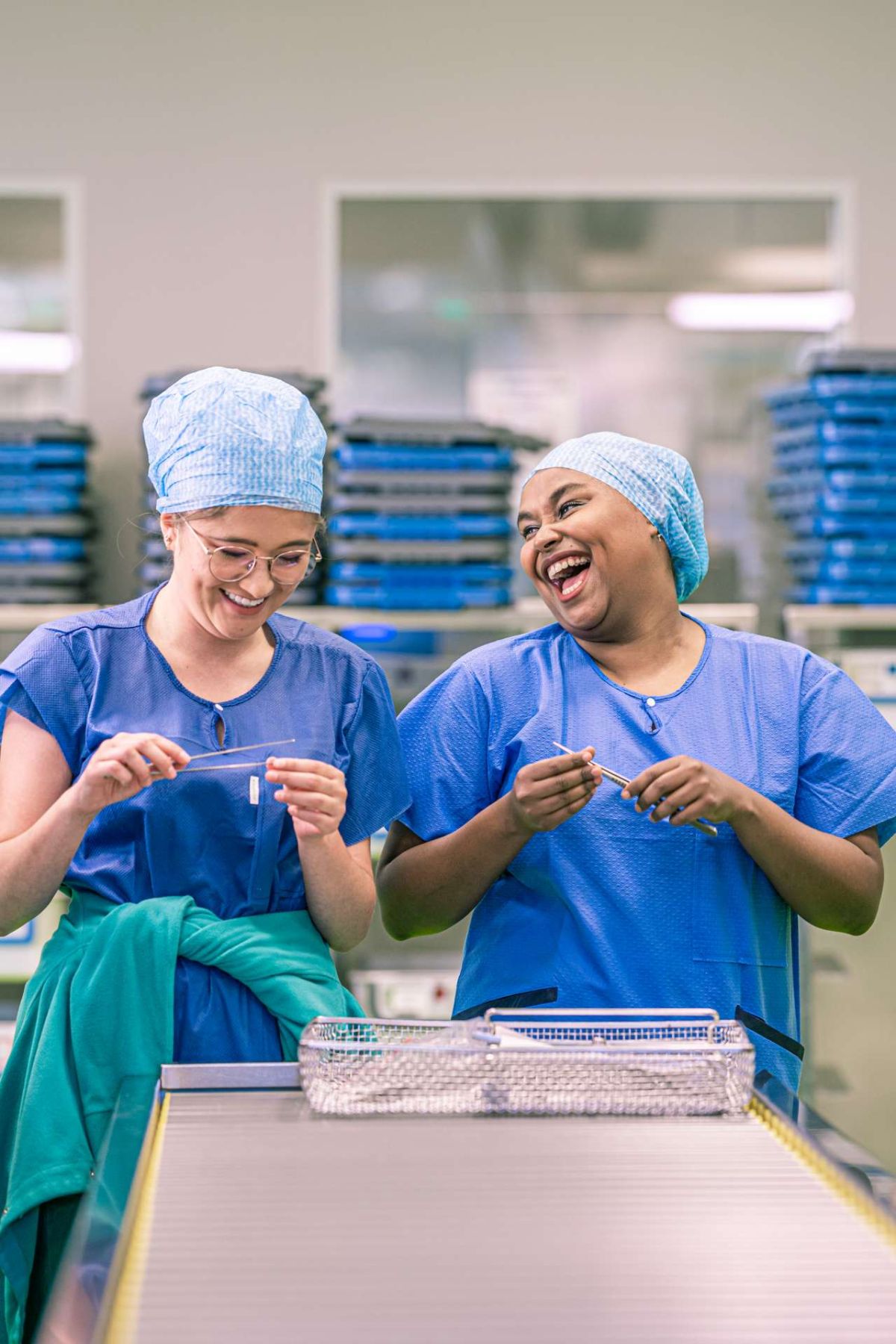 Zwei Medizinproduktetechnologinnen in blauer Berufskleidung und Haube stehen lachend an einem Arbeitstisch in der Aufbereitungseinheit für Medizinprodukte. Sie halten chirurgische Instrumente in der Hand und wirken engagiert und gut gelaunt.