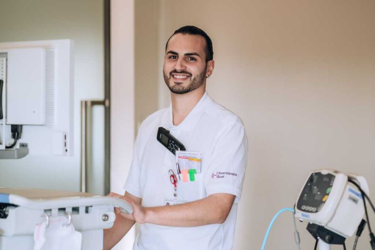 Ein junger Pfleger in weisser Arbeitskleidung des Universitätsspitals Basel steht lächelnd an einem medizinischen Arbeitsplatz mit Monitor und Geräten.