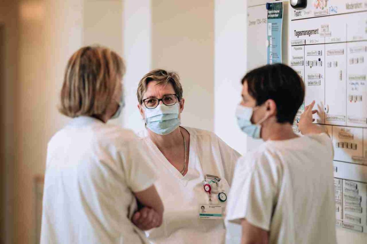 in Team von drei Frauen in weisser Berufskleidung und medizinischen Masken steht vor einem Wandboard mit Aufschriften wie 'Tagesmanagement'. Eine Frau, die in der Mitte steht, trägt eine Brille und einen Namensausweis und scheint aufmerksam zuzuhören. Eine andere zeigt mit einer Hand auf das Board, während die dritte Frau ihr den Rücken zuwendet. Die Szene zeigt eine konzentrierte Besprechung in einer professionellen Arbeitsumgebung.
